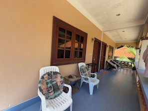 Un couloir abrité, avec deux chaises en plastique blanc, une petite table et une vue sur une maison avec un toit en tuiles.