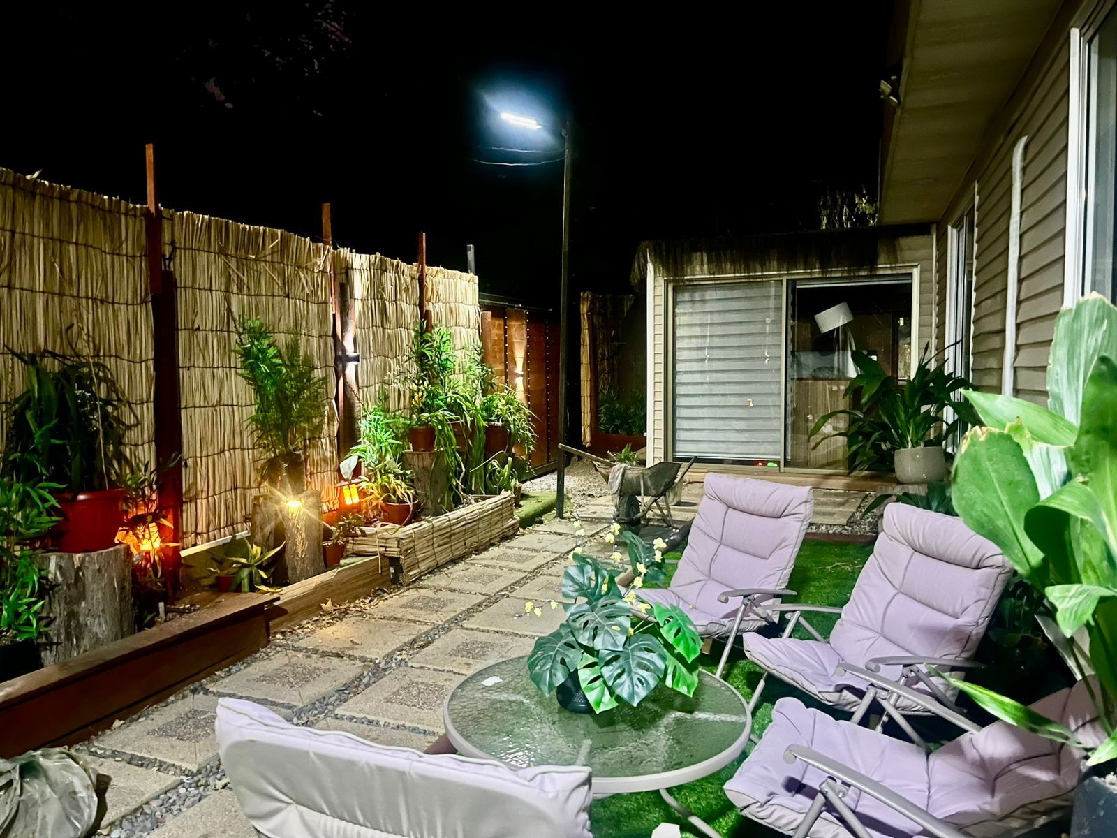 Un patio con mesa y sillas, rodeado de plantas en macetas y una cerca de bambú.
