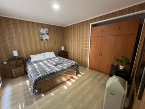 A bedroom with a wooden floor, a bed with a striped bedspread, a nightstand with a lamp, a wardrobe, and a potted plant. - Mountain Cabin with Private Hot Tub in San José de Maipo 0194A (El Manzano)