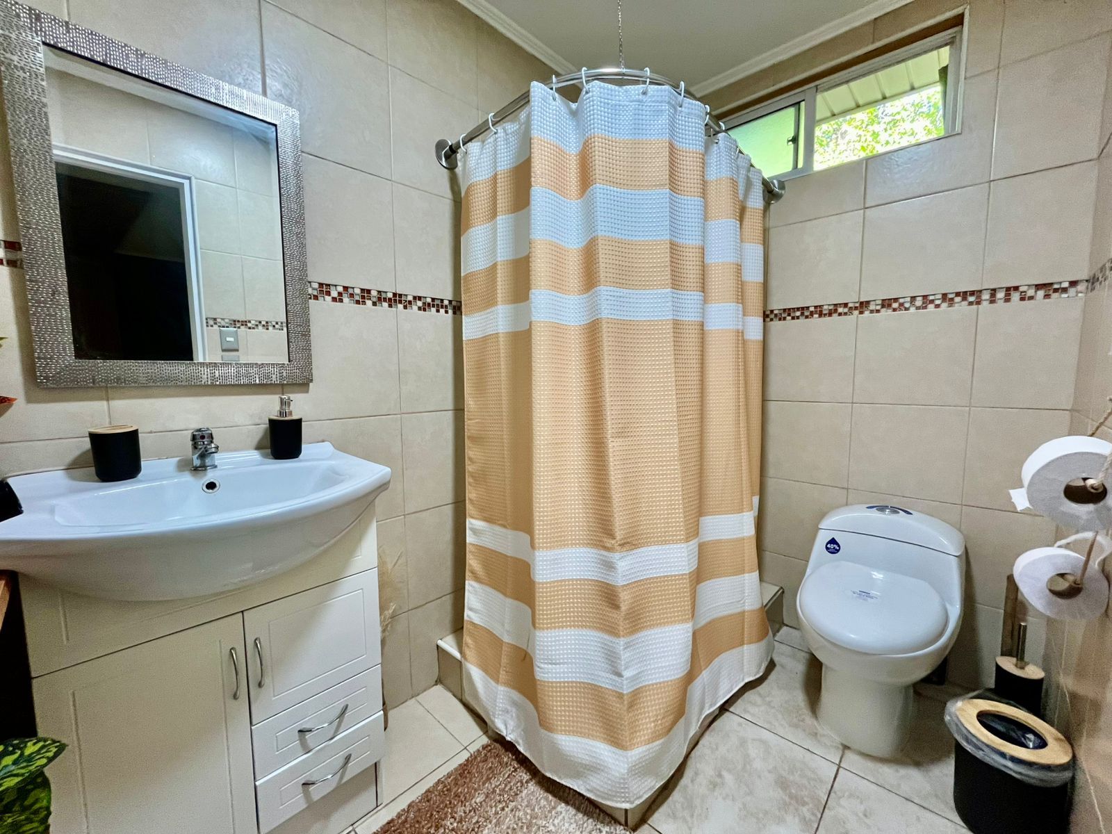Un baño con un lavamanos blanco, un espejo, una cortina de ducha a rayas, un inodoro y un pequeño cubo de basura.