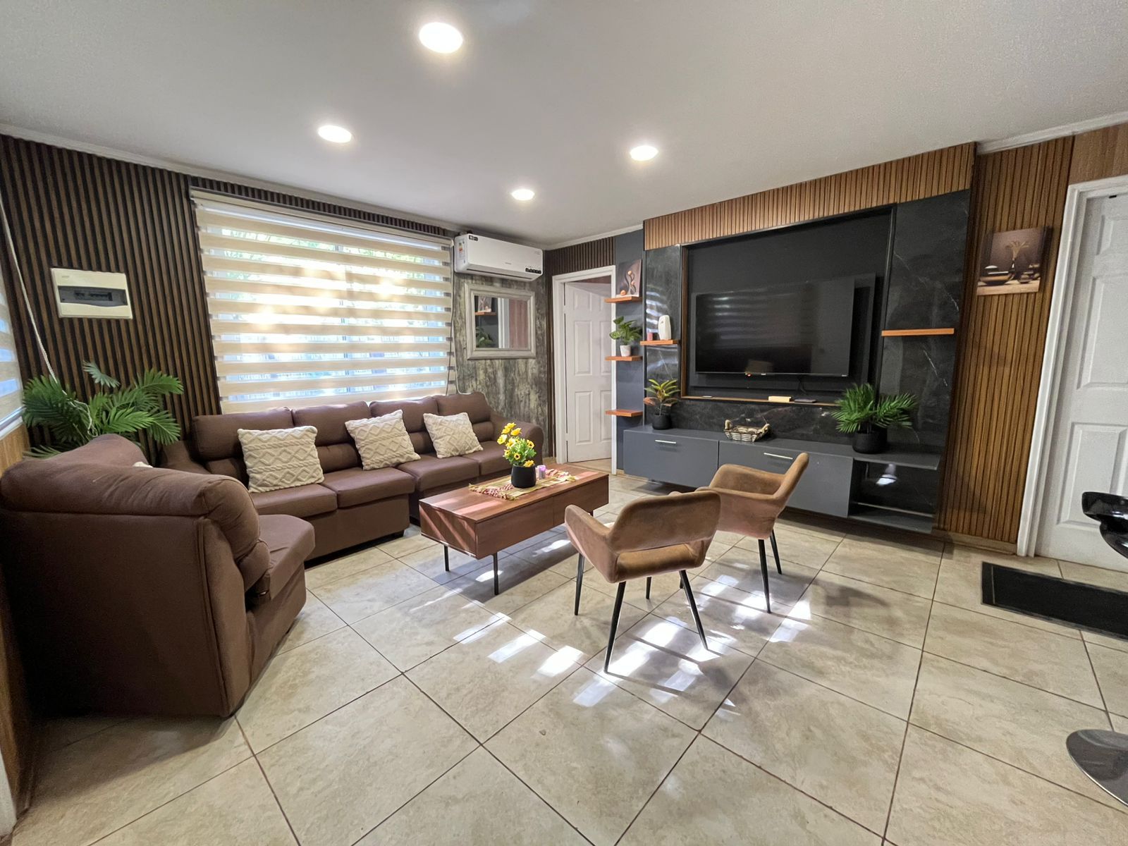 A modern living room with a sofa, coffee table, and chairs, a flat-screen TV, and a kitchen area with a countertop and cabinets.