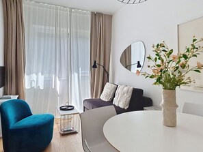 Una sala moderna con sofá, un sillón azul, una mesa redonda, una lámpara de pie y un florero con flores.