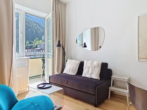 Una sala moderna con un sofá, una mesa de centro, una silla, una lámpara de pie y un ventanal que ofrece vistas a una zona boscosa.