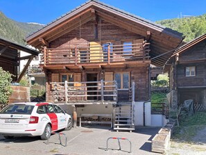 Xalet de fusta de dos pisos amb un balcó, un cotxe a la part davantera i un paisatge de muntanya de fons.