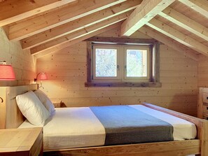 Ein Schlafzimmer im Dachgeschoss mit Holzbalken, geneigter Decke, einem Fenster mit Aussicht, einer Nachttischlampe und einem Bett mit Kissen.