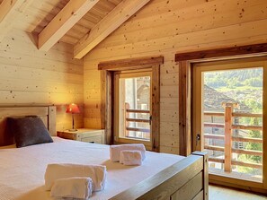 Una habitació de fusta amb un llit, una tauleta de nit, una làmpada i una finestra amb vista a un poble.