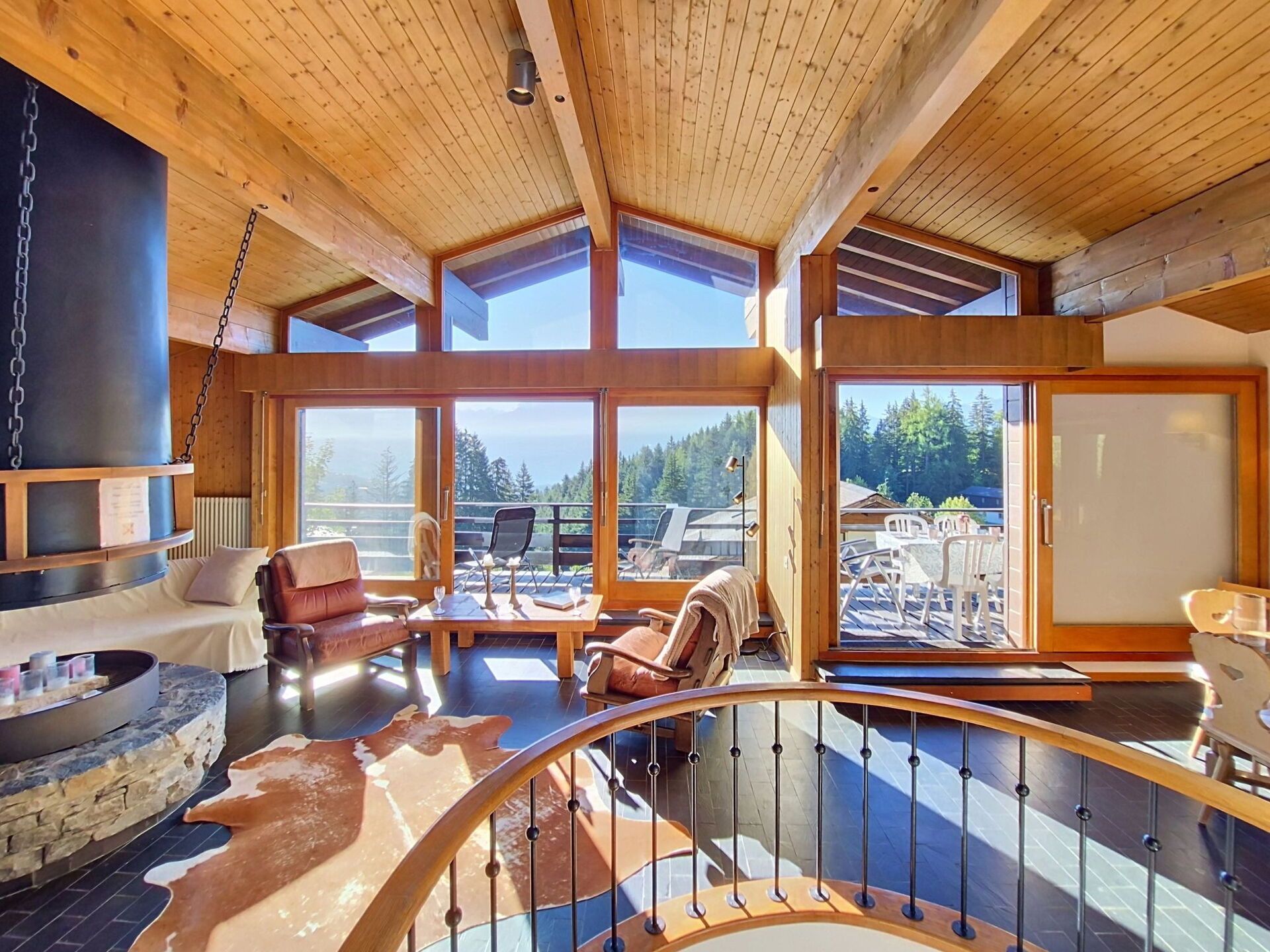 A spacious interior with a wooden ceiling, large windows offering a view of the outdoors, and a seating area with leather chairs and a stone fireplace.