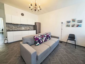 Ein modernes Wohnzimmer mit einem grauen Sofa, einer Chandelieber, einer Küche im Hintergrund und einem Sessel.