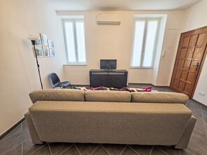 Ein Wohnzimmer mit einer Couch, einem Fernseher auf einem Schrank und zwei Fenstern.