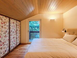 Un dormitorio con techo de madera, una cama con ropa de cama blanca, una puerta que da al exterior y una lámpara fijada a la pared.