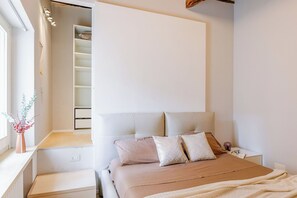 A bedroom with a bed, bedside tables, a nightstand, a window with curtains, and a built-in closet with shelves.