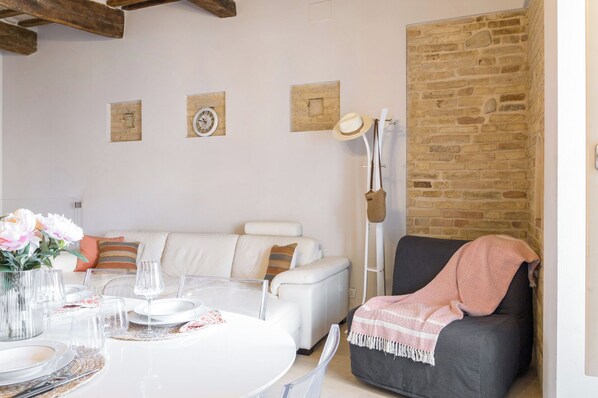 A living room with a white sofa, a dining table set with plates and glasses, a brick wall, and a hat hanging on a stand.