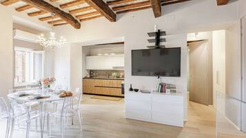Une cuisine moderne avec des poutres en bois, un coin repas avec des chaises transparentes et un téléviseur à écran plat fixé sur un meuble blanc.