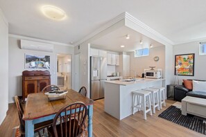 A modern kitchen with white cabinets, a central island with stools, and a dining area with a wooden table and chairs. - Sea and Salt - Palm Beach Coastal Escape (Gold Coast)