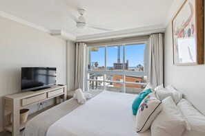 A hotel room with a bed, a flat-screen TV, a ceiling fan, a large window with a view of buildings and a crane, and a painting on the wall. - Sea and Salt - Palm Beach Coastal Escape (Gold Coast)