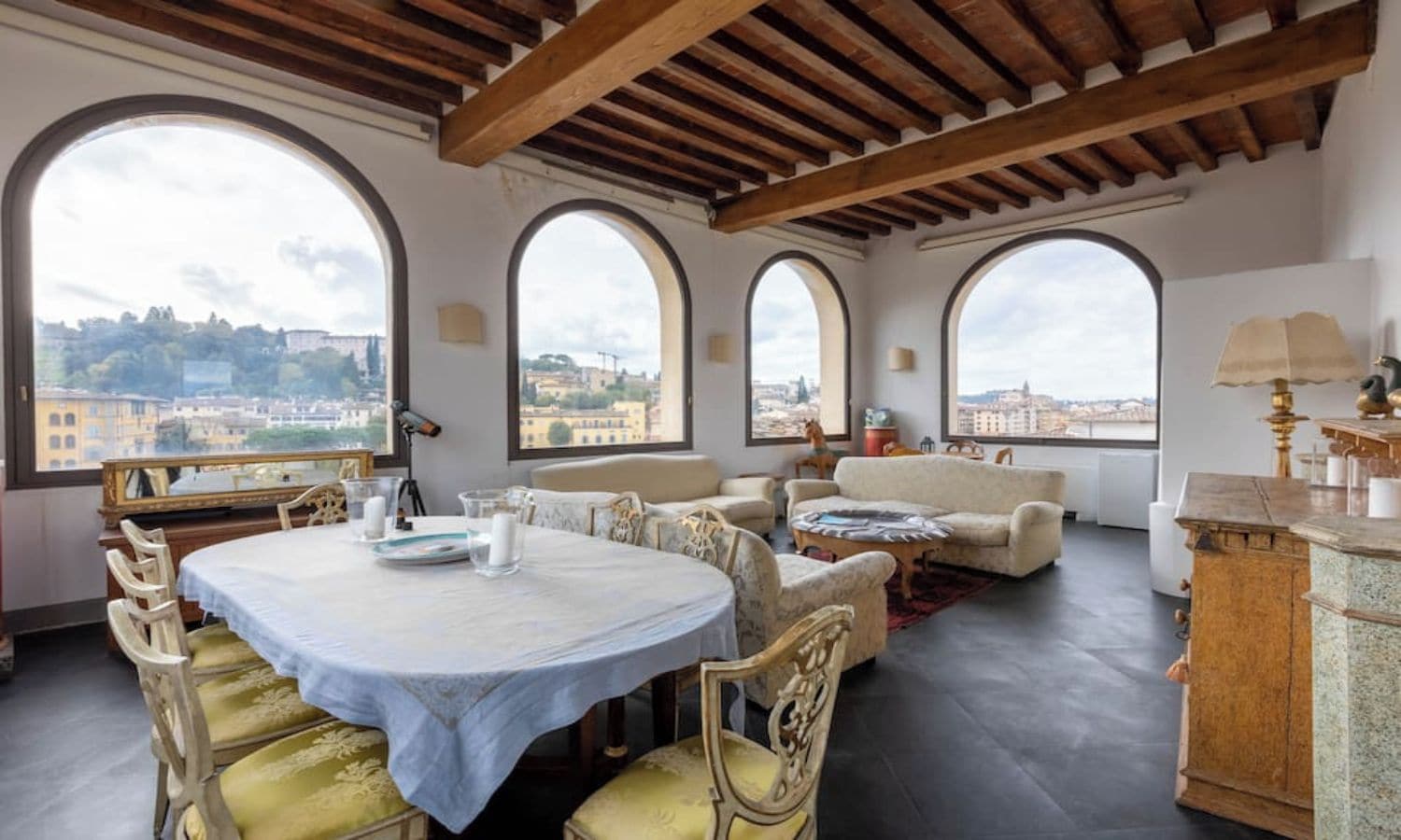 A dining area with a round table, chairs, and a view of the city through large arched windows.
