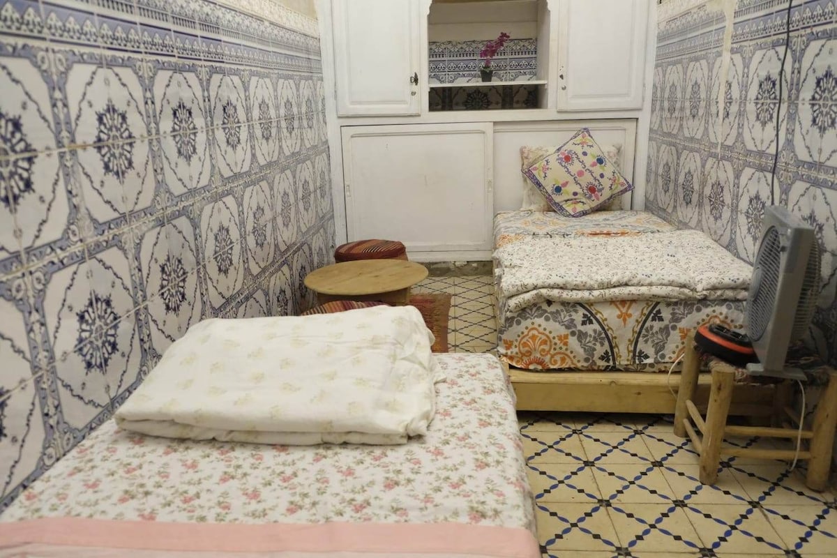 Una habitación pequeña y funcional con dos camas, una silla de madera, una mesita y un ventilador.