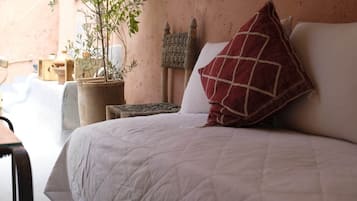 A bed with a white quilt, a red pillow, and a woven wall hanging. There is a potted plant to the left and a wooden stool.