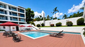Zona de piscina con sillones de descanso y sombrillas, rodeada por un edificio de varios pisos y fachada blanca.