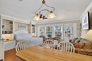 A well-lit room with a wooden dining table, white upholstered chairs, a sofa with floral upholstery, and a ceiling fan with three lights. - Tortuga Inn Beach Resort #219 | Direct Gulf & Beach Views (Bradenton Beach)
