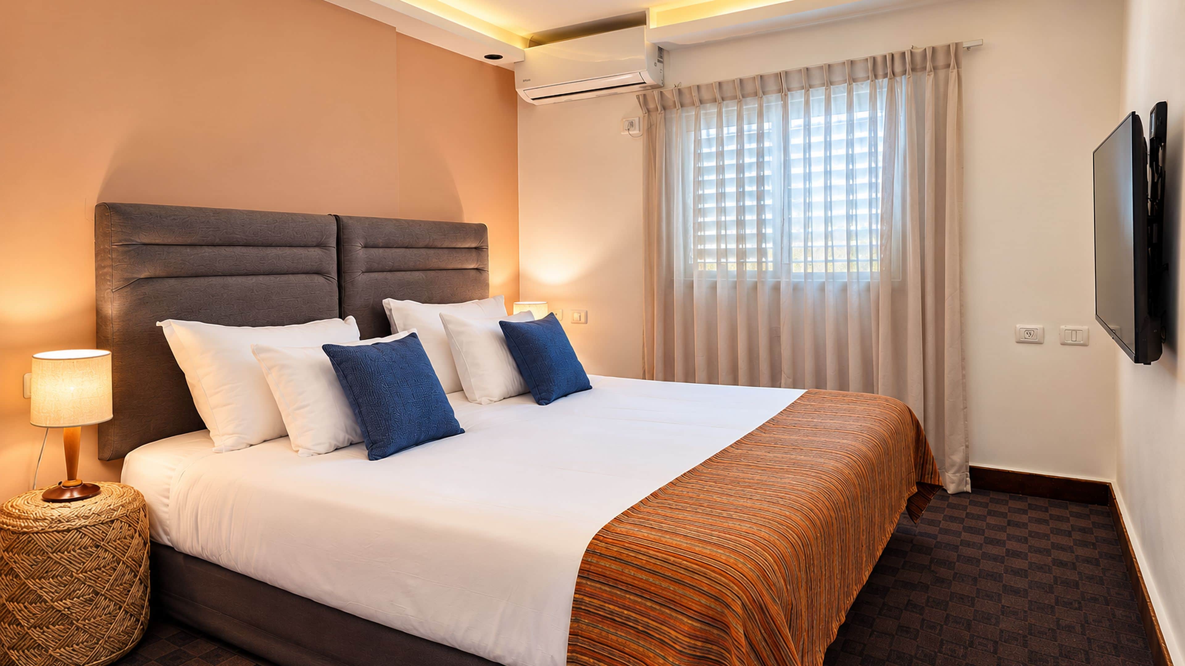 Una habitación de hotel con cama, televisor, aire acondicionado y ventana con cortinas.