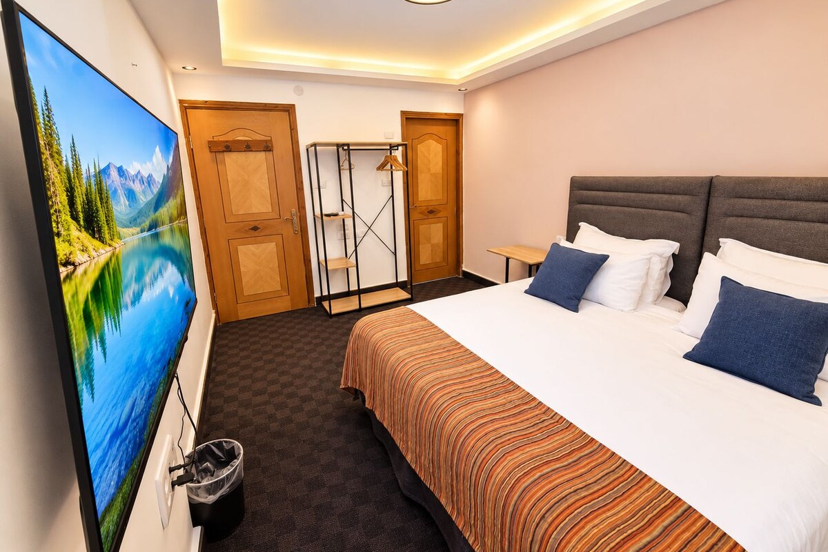 Una habitación de hotel con una cama grande, un televisor en la pared, un armario de madera y una mesita pequeña.