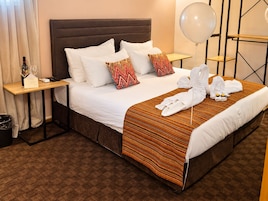 Habitación de hotel con cama, almohadas, mesita de noche con una botella y vasos, perchero con toallas y un globo sobre un soporte.