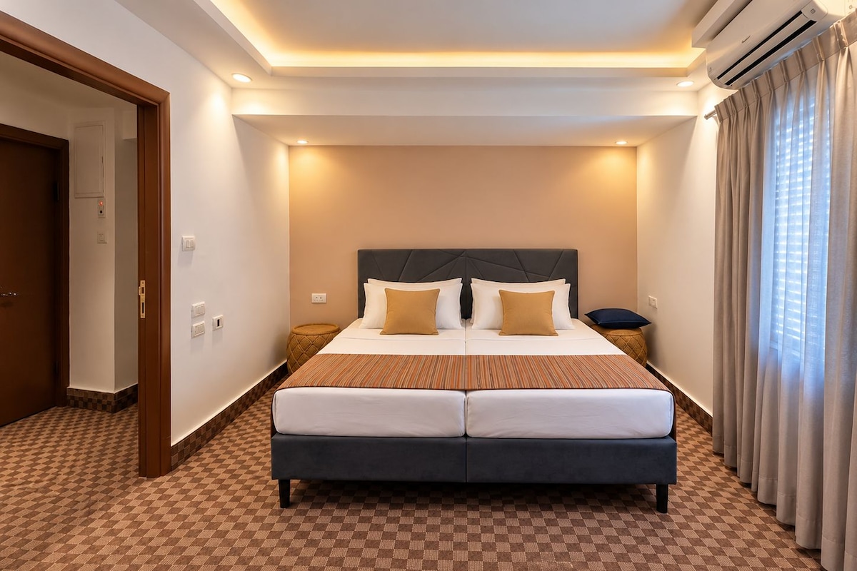 Una habitación de hotel con una cama grande, una mesita de noche, aire acondicionado instalado en la pared y una ventana con cortinas.