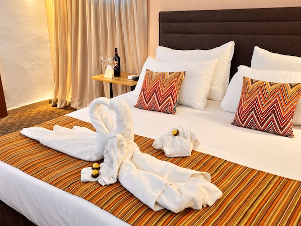 A hotel room with a bed, pillows, a bottle, glasses, and a towel arrangement.