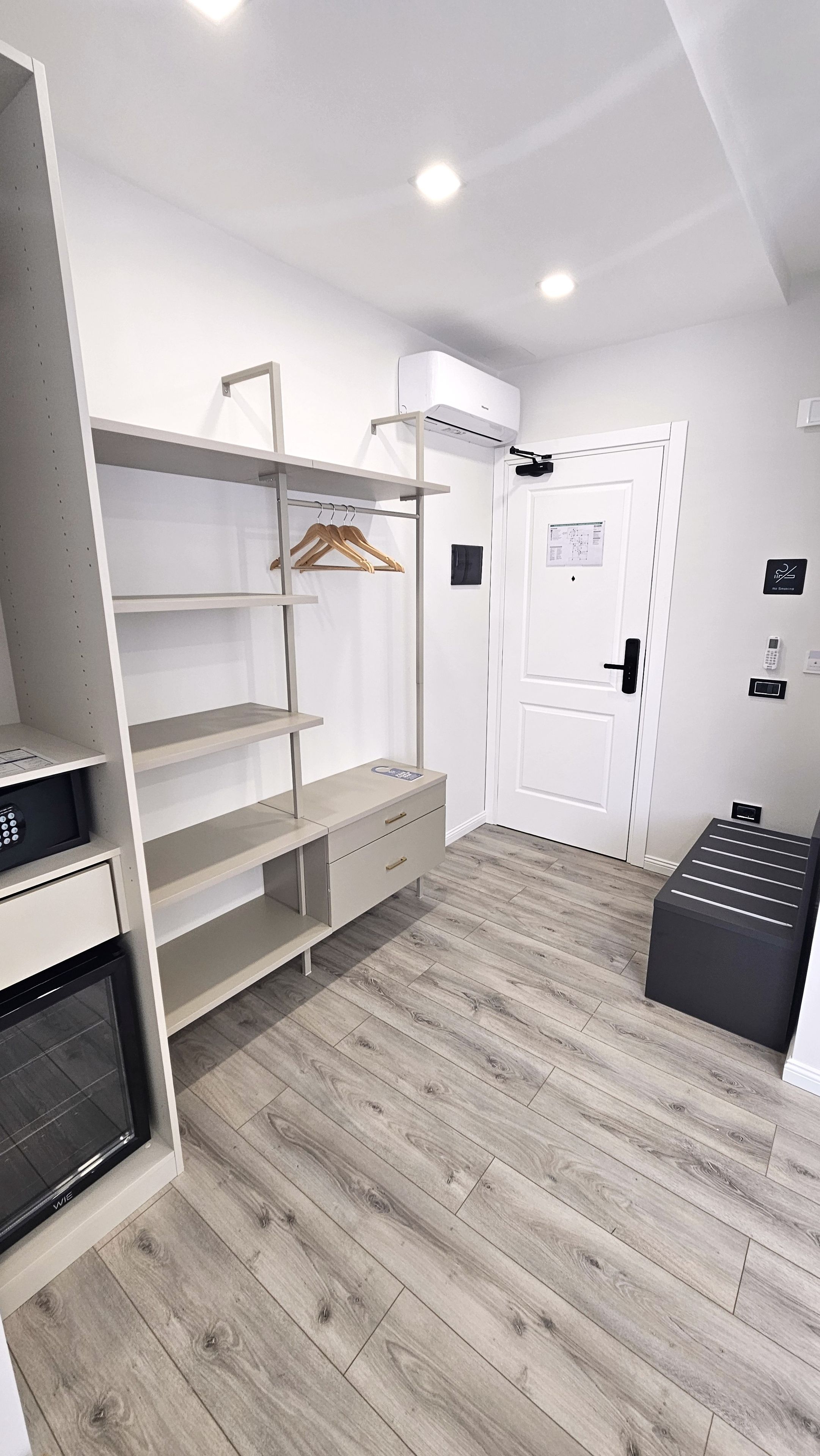A modern, well-lit room with a built-in oven, a wooden hanger rack, a white door with a keypad, and a black bench.