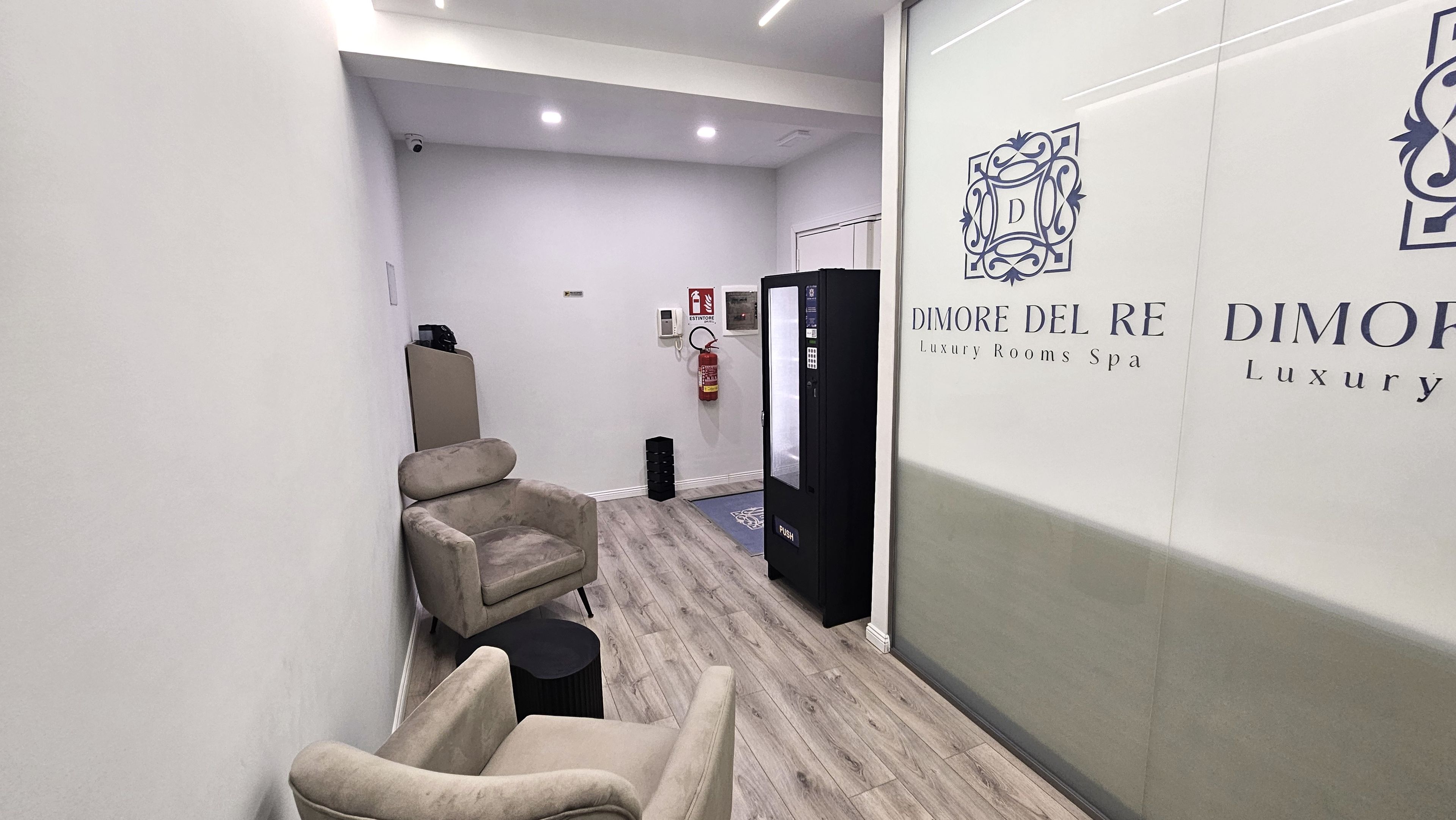 A hallway with a massage chair, a fire extinguisher, and a wall with a logo and text for 'DIMORE DEL RE Luxury Rooms Spa'.