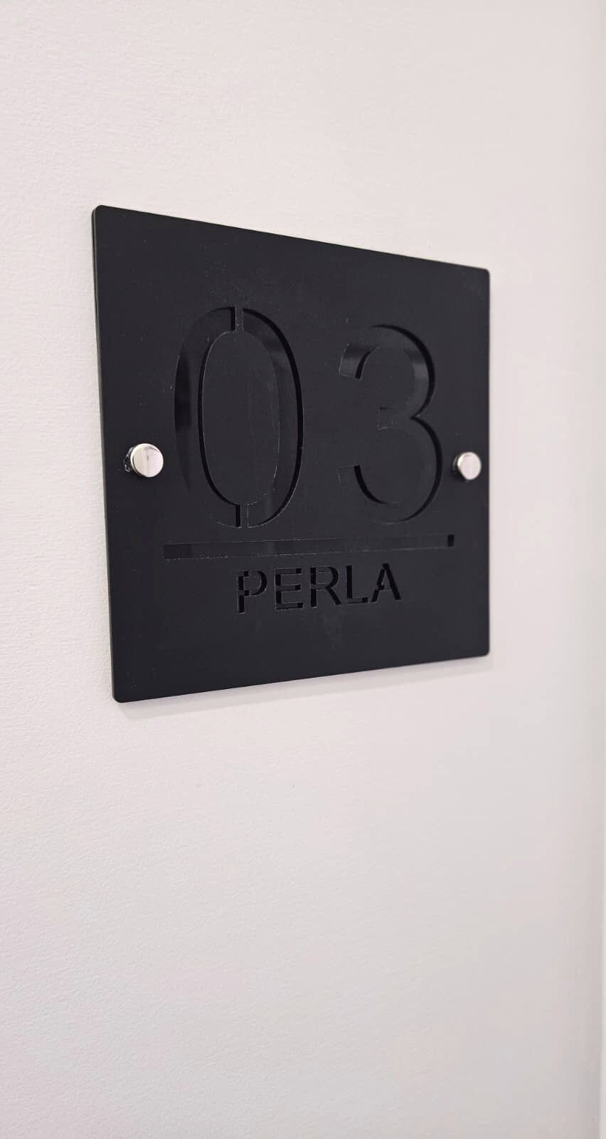 A black metal number plate with '03' and 'PERLA' engraved on it, mounted on a white wall.
