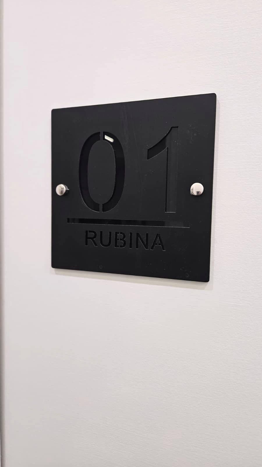 Une plaque en métal noir portant le numéro « 01 » et le mot « RUBINA », fixée à un mur blanc.