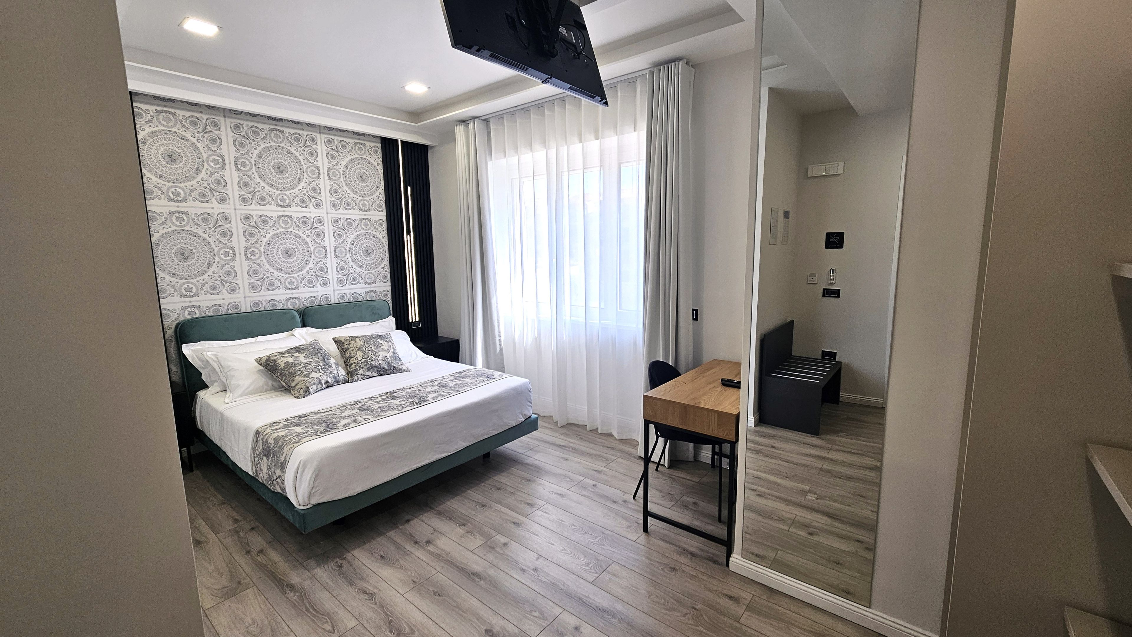 A modern hotel room with a bed, a desk, and a view of the city through large windows.