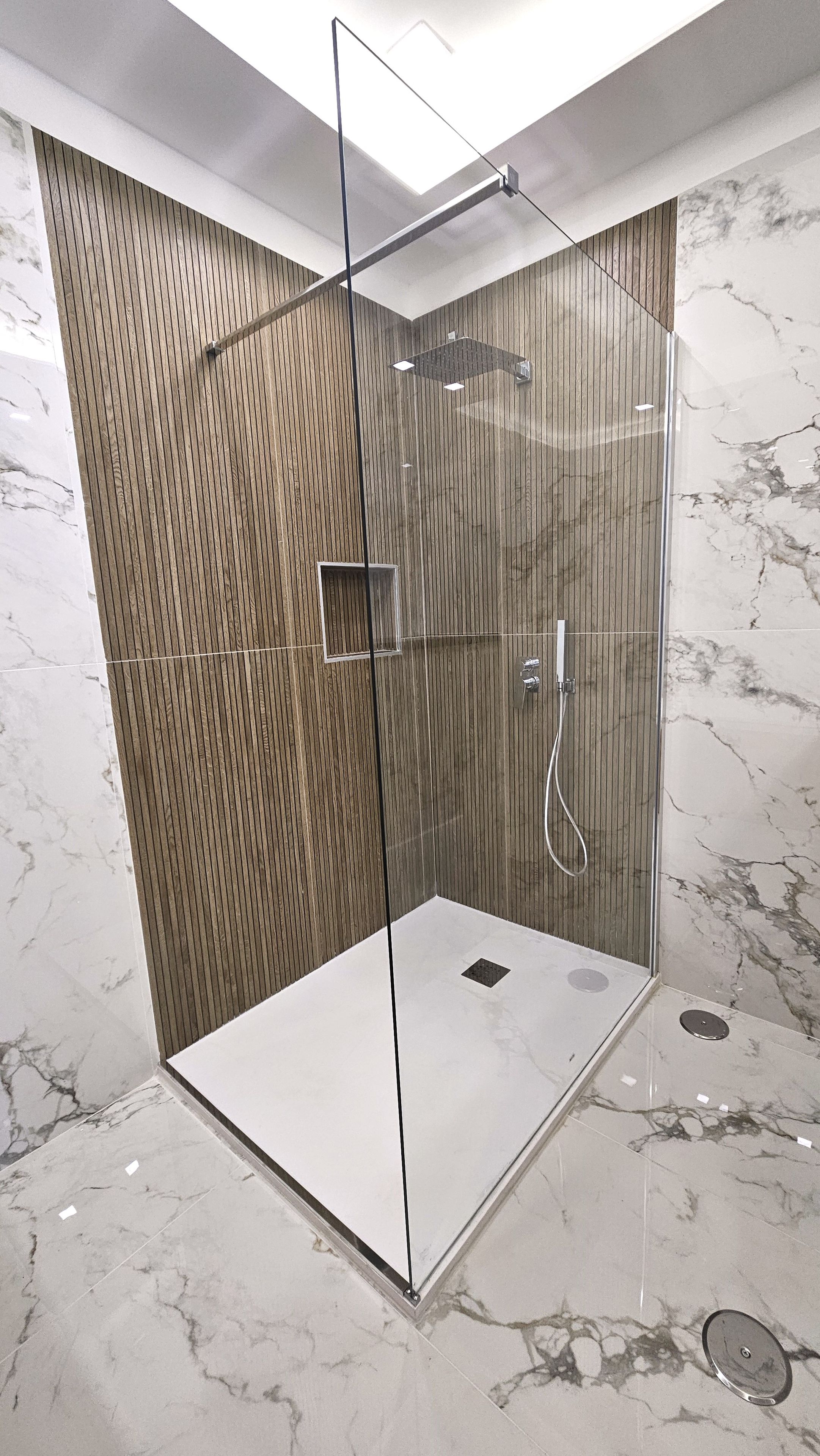Eine begehbare Dusche mit Glaswänden, einer Wand aus Holzpaneelen, einem Marmorboden und einer Deckenleuchte.