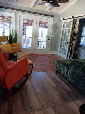 Un salon avec un plancher en bois, un canapé rouge, un canapé vert et un fauteuil jaune. Une grande fenêtre offre une vue sur l’extérieur.