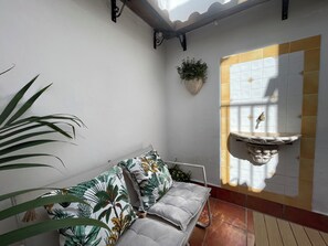 A room with a white wall, a window with a yellow frame, a hanging plant, a white sofa with a leaf pattern, and a small white sink. - Charming Little House (Requena)