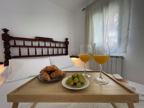 A wooden bed with a headboard, a bedside table with a tray holding croissants, grapes, and two glasses of orange juice.