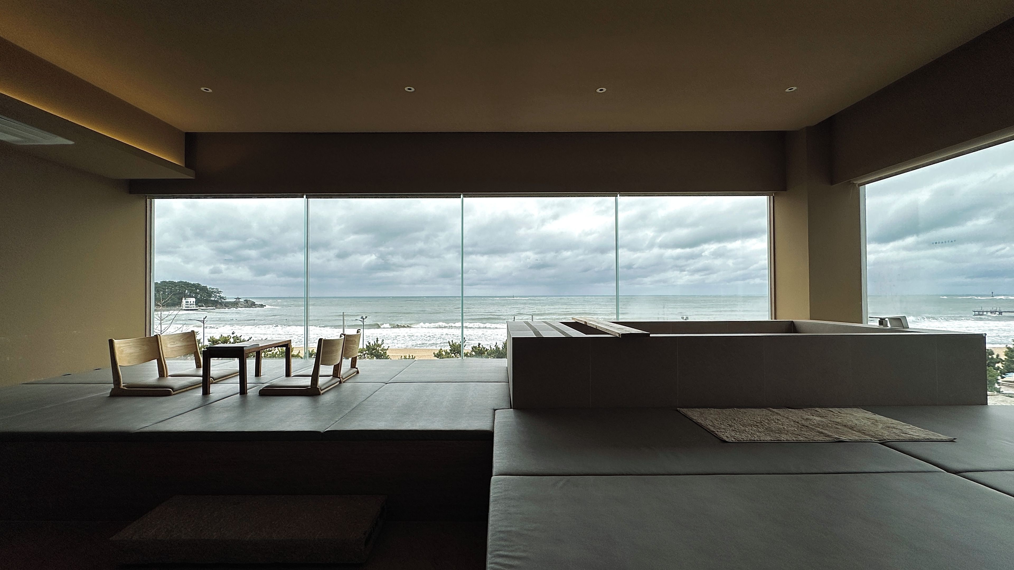 A modern interior with a large window overlooking the ocean, a seating area with cushions, and a minimalist design.