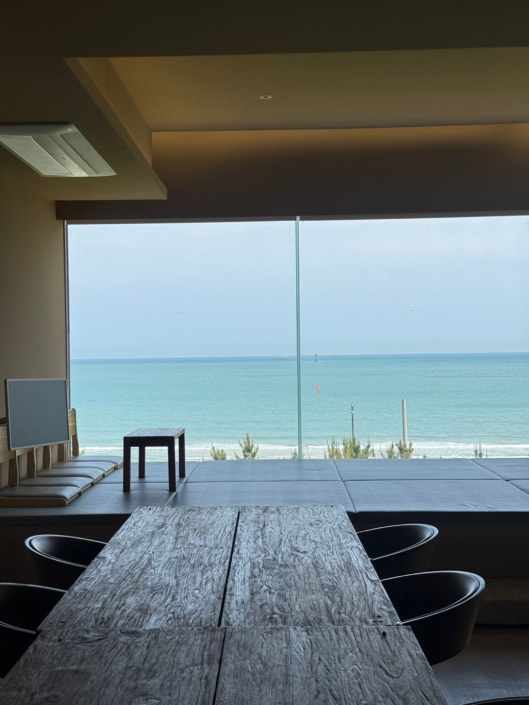 Une chambre moderne dotée d’une grande fenêtre donnant sur la mer, d’une table en bois et d’un coin salon avec des coussins.