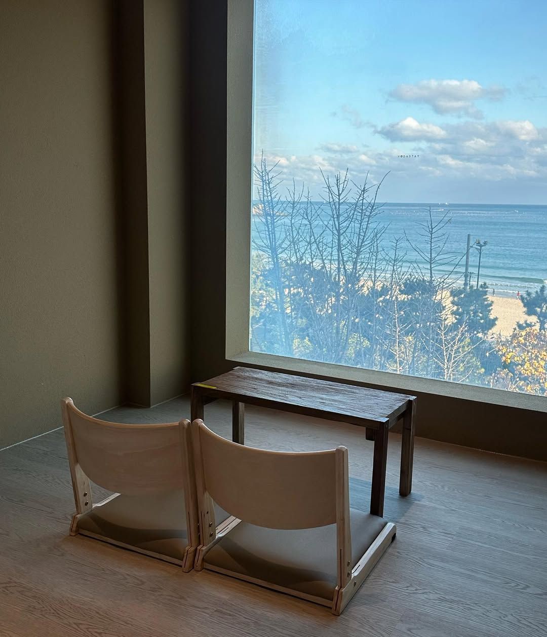 A room with a large window overlooking a beach and trees. There is a wooden bench and a small table.