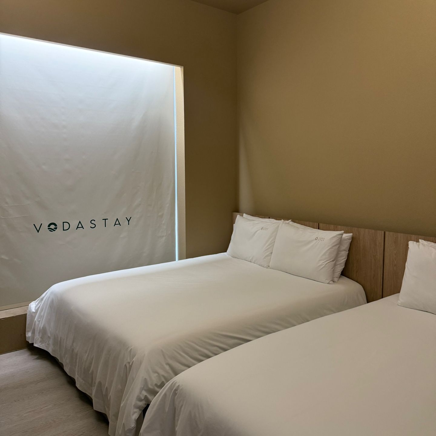 Une chambre d’hôtel avec deux lits, une tête de lit en bois et une grande fenêtre avec un store blanc.