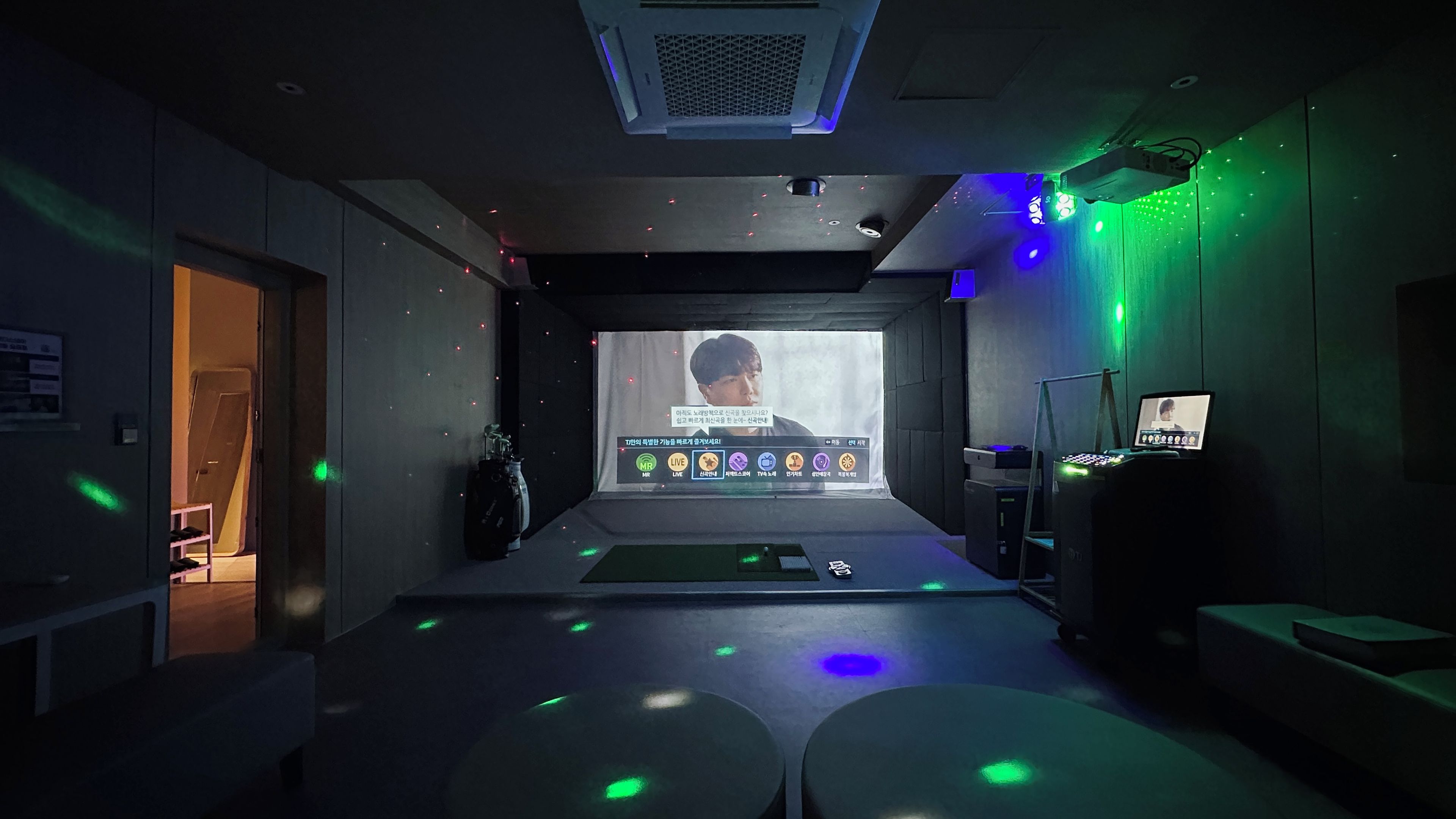 Een kamer met een projectiescherm waarop een persoon, een dj-installatie en een deur naar een andere ruimte te zien is.
