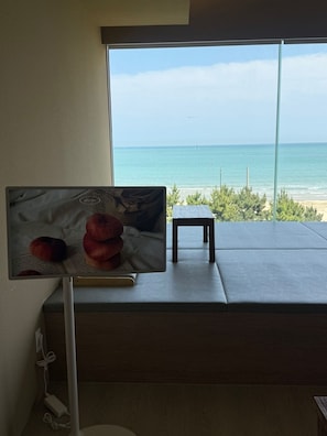 A room with a large window overlooking a beach, a small table, and a wall-mounted television displaying an image of apples. - VODASTAY Songjeong (Busan)