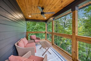 A covered outdoor seating area with wicker furniture and a table, overlooking a forested landscape. - Modern Mountain Chalet | Views, Hot Tub & Fire Pit (Gatlinburg)