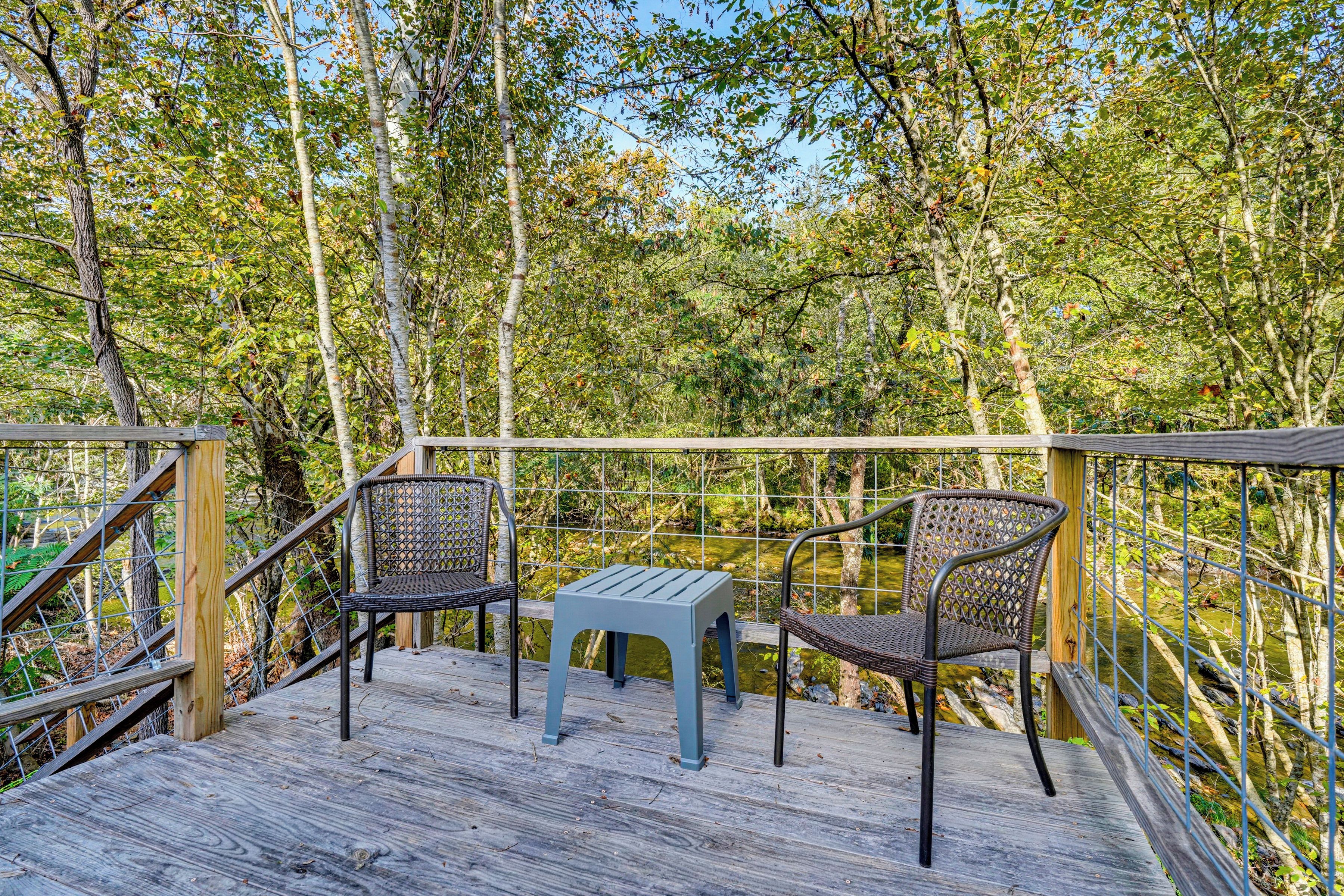 A wooden deck with two wicker chairs and a small table, surrounded by a metal railing and dense foliage.