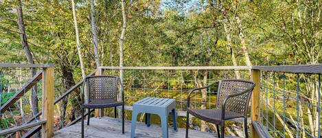 A wooden deck with two wicker chairs and a small table, surrounded by a metal railing and dense foliage.
