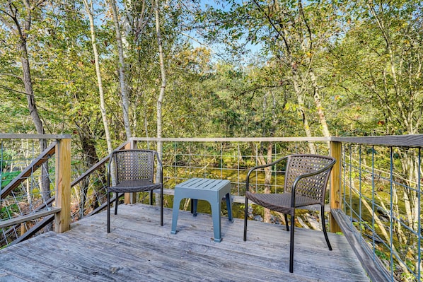 A wooden deck with two wicker chairs and a small table, surrounded by a metal railing and dense foliage. - Luxury River Retreat with Hot Tub, Fire Pit & Fly Fishing, private river deck (Sevierville)