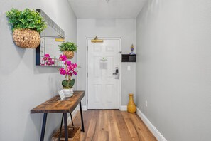 A hallway with a wooden bench, a potted orchid, a wicker basket, a white door with a fire evacuation plan, and a small shelf with decorative items. - 4505 Minutes to Magic! 3/3 Resort Condo Near Disney (Orlando)