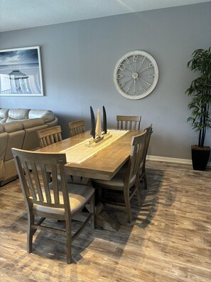 A dining area with a wooden table and chairs, a clock on the wall, a sofa, and a potted plant. - 1304 Spacious 3/3 Resort Condo Near Disney (Orlando)